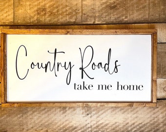 May include: A white wooden sign with a brown frame, featuring the text "Country Roads take me home" in black cursive font.