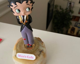 Può includere: Una statuetta in ceramica di Betty Boop vestita con un abito nero con cravatta dorata e un cappello a cilindro dorato. La statuetta è in piedi su una base di marmo bianco con glitter dorato e un cartello rosa che recita "Max's Cafe".