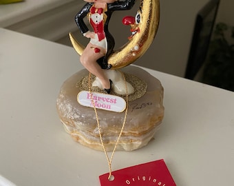 Può includere: Una statuetta di Betty Boop vestita con uno smoking, seduta su una luna crescente con una finitura dorata. La luna ha un viso e tiene un cappello a cilindro. La statuetta è su una base di marmo bianco e marrone con le parole "Harvest Moon" e "Ron Lee '03" scritte su di essa. La statuetta ha un'etichetta che dice "The Original Ron Lee Collection".