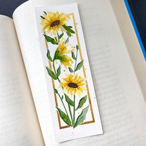 May include: Watercolor bookmark featuring yellow sunflowers with green leaves and a gold border. The bookmark is rectangular and is placed inside an open book.