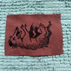 Może przedstawiać: Prostokątna, brązowa naszywka z czarną ilustracją wyjącego wilka z krzyżykiem nad okiem. Wilk leży na plecach z łapami w górze. Naszywka jest umieszczona na teksturowanym, jasnoniebieskim tle.