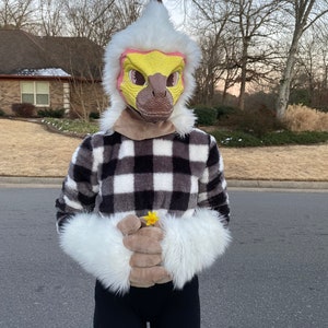 May include: A person wearing a bird-like mask with a yellow face, brown beak, and white feathered head. They are wearing a black and white checkered fleece jacket, black leggings, and white furry gloves. A small yellow flower is held in the gloves.