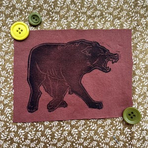 May include: A maroon fabric square featuring a black linocut print of a snarling bear. The image is set against a patterned background with yellow and green buttons.