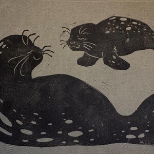 Puede incluir: Una impresión en tinta negra sobre tela de color beige claro que muestra dos focas moteadas. La foca más grande está situada en la parte inferior, mirando hacia arriba a una foca más pequeña que nada encima. Ambas focas tienen bigotes.