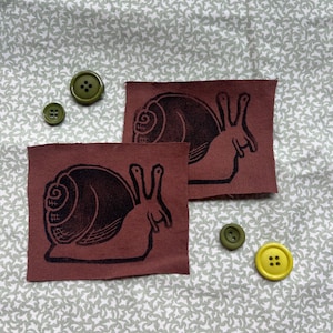 May include: Two brown fabric patches with black snail illustrations, accompanied by green and yellow buttons. The patches are rectangular and have a rough, textured appearance. The buttons vary in size and color, adding a playful contrast to the design.