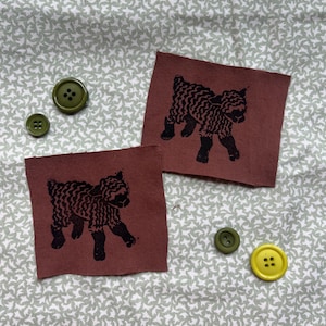 Könnte beinhalten: Zwei braune Stoffflicken mit schwarzen Lamm-Illustrationen, zusammen mit drei Knöpfen in Grün- und Gelbtönen. Der Hintergrund ist ein hellgrüner Stoff mit weißem Blumenmuster. Geeignet für Handarbeiten.