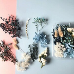 May include: Assortment of dried flowers and foliage in shades of blue and white, including hydrangeas, baby's breath, and sprigs of greenery. The flowers are arranged in a clear plastic container.
