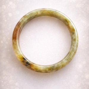 Puede incluir: Un brazalete de jade redondo. El brazalete es una mezcla de colores, incluyendo verde, marrón y amarillo. El brazalete es liso y pulido, con una apariencia natural y orgánica. El fondo es blanco suave y borroso.