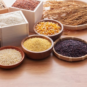May include: An assortment of grains and seeds are presented in wooden containers and bowls. The image features white rice, red rice, corn, and other grains, highlighting their textures and colours. The arrangement suggests a focus on healthy eating and natural foods.