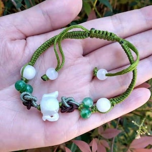 Könnte beinhalten: Ein grünes geflochtenes Armband mit weißen und grünen Perlen und einer weißen geschnitzten Figur. Das Armband wird in einer Hand gehalten, mit einem verschwommenen Hintergrund aus grünem und rosa Laub.