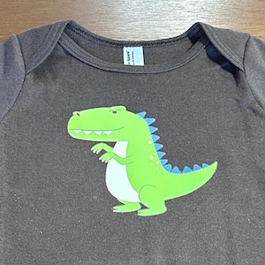 May include: A brown baby bodysuit with a green dinosaur graphic. The dinosaur has a white belly and blue spikes. The bodysuit has short sleeves and three snaps at the bottom.