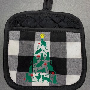 May include: Black and white checkered pot holder with a black quilted border and hanging loop. The center features a green cat-shaped Christmas tree design with paw prints and fish bones. A fun and functional kitchen accessory.
