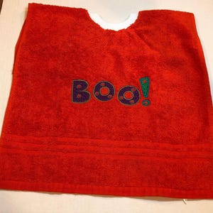 May include: A bright red terry cloth bib with a white neckline. The word "BOO!" is embroidered in purple and green. The bib is designed to protect clothing during mealtime or other activities.
