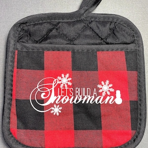 May include: Black and red buffalo plaid pot holder with a loop for hanging. The pot holder features the text "Let's Build a Snowman" in white script, along with snowflake and snowman graphics. A festive kitchen accessory.