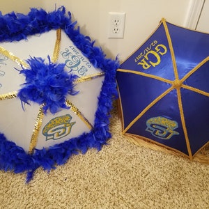 May include: Two white and blue umbrellas with gold trim and a blue and gold logo. The umbrellas are decorated with blue feathers. One umbrella has the text "GCR 02/11/2017" and the other has the text "When Love Begins".