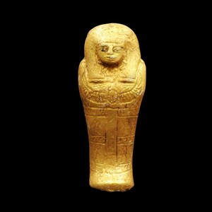 May include: A small, golden-colored ancient Egyptian ushabti figurine. The figure has a detailed face, a headdress, and intricate carvings on the body. The ushabti is standing upright, with arms crossed, and is set against a black background.