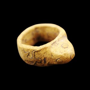 May include: A tan-colored ring with a rounded shape and a smooth texture. The ring has dark markings and a slightly bulbous design on one side. The background is solid black.