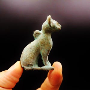 Peut inclure: Petite figurine de chat en bronze vieilli, en position assise. La sculpture présente une patine brun-verdâtre vieillie, suggérant l'âge. Les oreilles du chat sont dressées et il semble regarder sur le côté. La figurine est tenue sur un fond sombre.