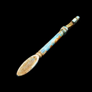 Può includere: Un utensile lungo e sottile, in stile antico, con un manico azzurro e un'estremità arrotondata di forma ovale. Il manico presenta dettagli marroni e dorati. L'oggetto è posto su uno sfondo nero.