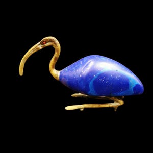 May include: A bird figurine featuring a smooth, deep blue body with subtle white and light blue veining. The head, long curved beak, and legs are crafted from a tan, bone-like material, with a small, bright red circular eye detail.