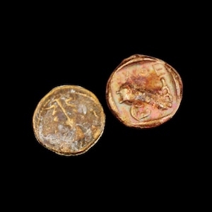 Puede incluir: Dos monedas antiguas de oro con desgaste visible. Una moneda presenta una imagen detallada de una figura, mientras que la otra muestra un diseño diferente. Las monedas están sobre un fondo negro.
