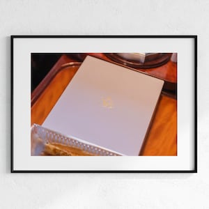 Puede incluir: Un objeto plano, rectangular y de color claro con una superficie lisa y un emblema dorado centrado. El objeto está colocado en ángulo, mostrando su panel superior limpio y de color sólido, junto con bordes afilados y definidos.