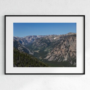 May include: A framed landscape photograph of a mountain valley under a clear blue sky. The valley is filled with green trees and the mountains have snow-capped peaks. The photograph is in a black frame.