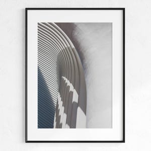 May include: Framed architectural print featuring a close-up of a modern building's curved facade. The image showcases repeating, parallel lines and shadows, creating a play of light and texture. The color palette is primarily neutral, with shades of gray and white.