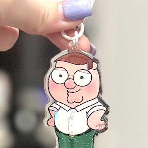 May include: A clear acrylic keyring featuring a cartoon character with brown hair, glasses, and a white shirt. The keyring has a white clasp and a glittery finish. The character is wearing green trousers and brown shoes.