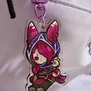 May include: A clear acrylic charm featuring a cartoon character with pink and purple accents. The charm is attached to a purple metal clasp. The character is wearing a hooded outfit.
