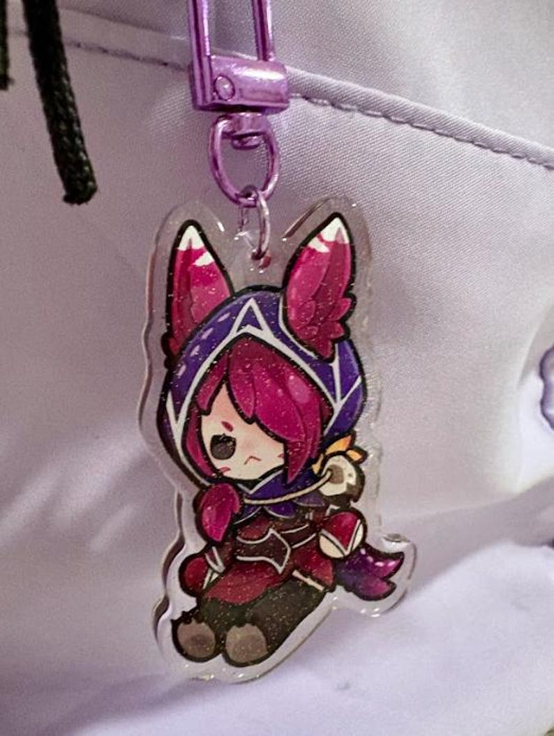 May include: A clear acrylic charm featuring a cartoon character with pink and purple accents. The charm is attached to a purple metal clasp. The character is wearing a hooded outfit.