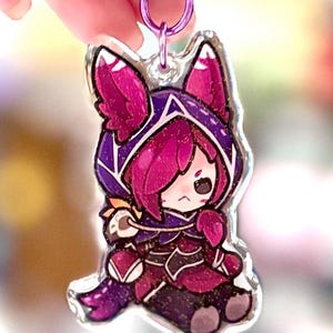 May include: A clear acrylic keychain featuring a cartoon character in a purple and magenta outfit with a hood. The keychain has a silver-colored clasp and a purple strap. The character has pink hair and a slightly sad expression.