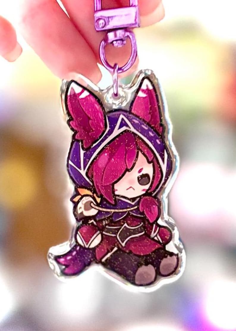 May include: A clear acrylic keychain featuring a cartoon character in a purple and magenta outfit with a hood. The keychain has a silver-colored clasp and a purple strap. The character has pink hair and a slightly sad expression.