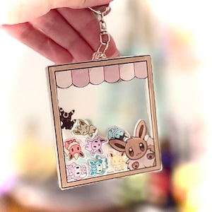 May include: A clear, square keyring with a light brown border and a pink and white awning design at the top. Inside are various cartoon characters in different colours, including a brown bunny-like character. The keyring is held by a silver chain.