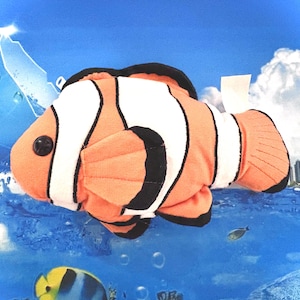 Könnte beinhalten: Ein Plüschspielzeug eines Clownfisches mit orangefarbenen, weißen und schwarzen Streifen. Das Stofftier hat ein Knopfauge und befindet sich vor einem blauen Hintergrund mit anderen Fischen und einer Stadtlandschaft.