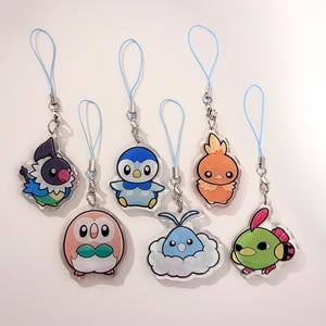 May include: A collection of seven cartoon character charms with blue lanyards. The charms feature various colorful designs, including a penguin, an owl, and a bird-like creature. Each charm is attached to a silver-colored clasp.