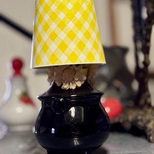 May include: A small, black, glossy lamp with a yellow and white checkered lampshade. The lamp has a round base and a decorative floral element. The lamp is on a gray surface.