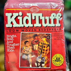 May include: A pink Kid Tuff loom woven bedspread in plastic packaging. The red label features the brand name in large white letters, a photo of three children, and text that reads "Bright. Bold. And Tougher Than Kids."