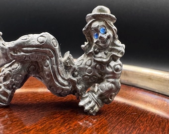 May include: A silver-colored clown figurine with blue gem eyes and a small hat. The clown is in a reclining pose, with a detailed design on the costume. The figurine is on a wooden surface.