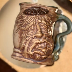 May include: A ceramic mug with a brown and tan face sculpted on the front. The face has a furrowed brow, a large nose, and a wide mouth. The mug has a green handle.