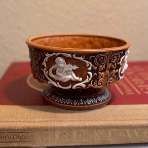 May include: A small, brown ceramic bowl with a white cherub design. The bowl has a raised base and intricate floral details.