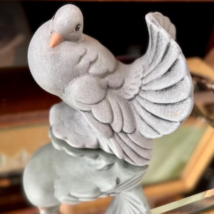 May include: A gray ceramic dove figurine with detailed feather textures and a peach-colored beak. The dove is perched on a reflective surface, creating a mirrored effect. The figurine is a decorative item, likely for display.