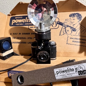 May include: A vintage black and silver camera with a flash attachment and a box labeled "Improved Powellite - Portable Sunshine". The box also has the text "Unconditionally Guaranteed" and a drawing of a person using the flash. The camera is connected to a black cord and a black and brown flash unit.