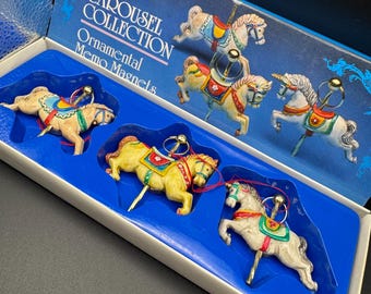 Puede incluir: Un conjunto de imanes de memo de caballos de carrusel en una caja azul. La caja dice "The Carousel Collection Ornamental Memo Magnets". Los imanes son coloridos, con detalles como sillas de montar y riendas.