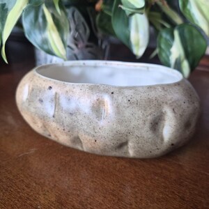 May include: A low, oval ceramic bowl with a speckled beige and tan glaze. The exterior surface features a series of vertical indentations around the perimeter, while the interior is finished in a smooth, plain white glaze.
