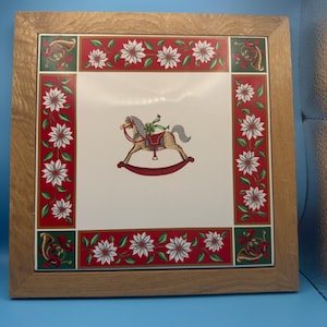 May include: Square wooden framed decorative tile with a Christmas theme. The center features a rocking horse illustration. The border has a red background with white flowers and green accents. The frame is made of light brown wood.