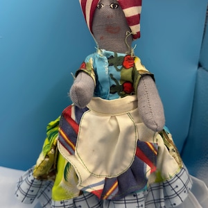 May include: Handmade cloth doll with a gray head and arms, wearing a colorful patchwork dress and a striped head wrap. The doll has a white apron and simple facial features. A unique, handcrafted item.