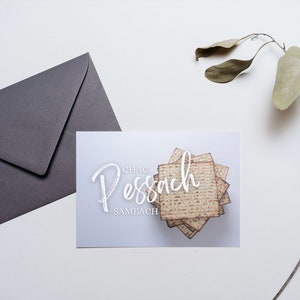 Könnte beinhalten: Eine weiße Karte mit dem Text "Chag Pesach Sameach" in weißer Schrift. Ein Stapel Matze ist in der Mitte der Karte.