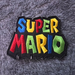May include: A black embroidered patch with the words "SUPER MARIO" in colorful letters. The letters are outlined in black and have a jagged edge.