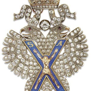 Op de afbeelding: Een gedetailleerde broche met een dubbelkoppige adelaar, bezet met talrijke diamanten. De vleugels van de adelaar zijn uitgespreid, en een blauw geëmailleerd kruis met een gouden figuur staat centraal. Een met diamanten bezette kroon staat boven de adelaarskoppen.